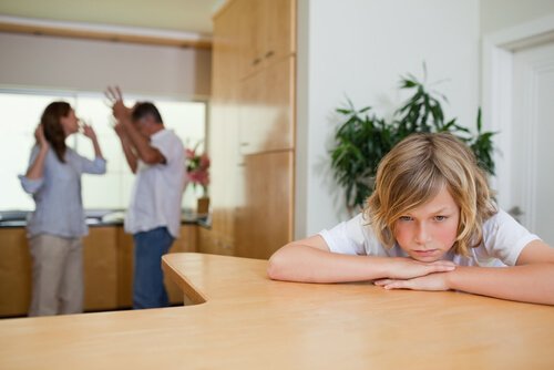 Chłopiec siedzący z głową leżącą na stole opartą na rękach, w tle kłótnia rodziców
