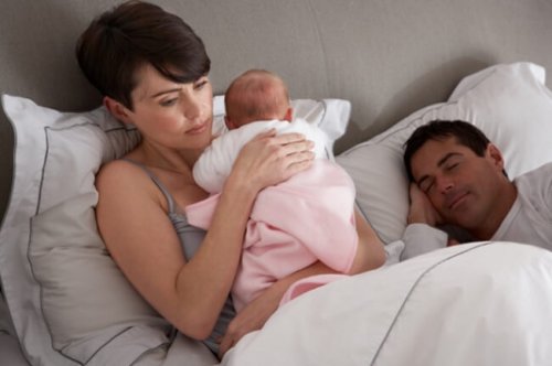 Mama przytulająca niemowlę w łóżku, obok śpiący tata