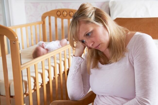 Smutna mama podpierająca głowę na ręku, w tle kojec z dzieckiem