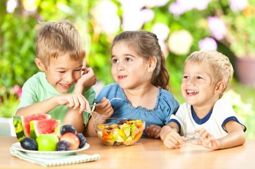 Dwóch chłopców i dziewczynka siedzący przy stole i jedzący zdrowe przekąski dla dzieci - owoce z miski