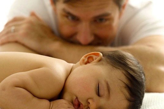 Tata z głową opartą na rękach przyglądający się śpiącemu dziecku