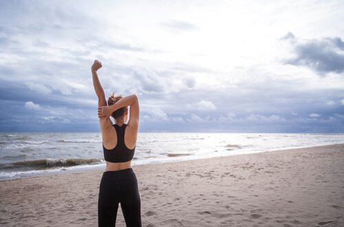 Kobieta w sportowym stroju stojąca tyłem i rozciągająca się na plaży