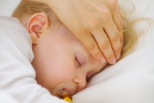 Mama przykładająca dłoń do czoła dziecka śpiącego ze smoczkiem w ustach