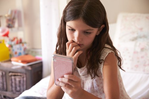 Dziewczynka siedząca na łóżku, wpatrzona w telefon i gryząca paznokcie