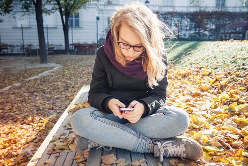 Nastolatka siedząca po turecku na ławce w jesiennym parku, pisząca na telefonie