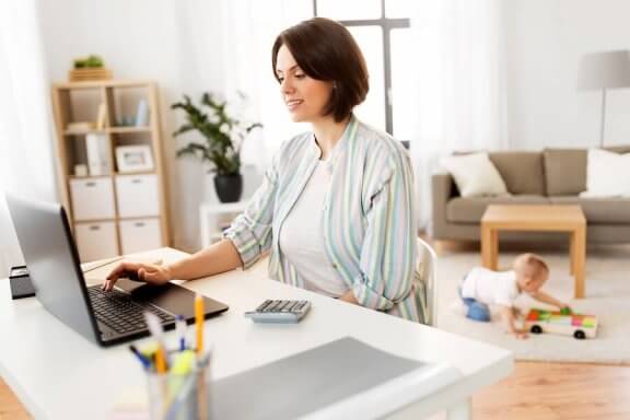 Mama siedząca przy laptopie, w tle bawiące się dziecko